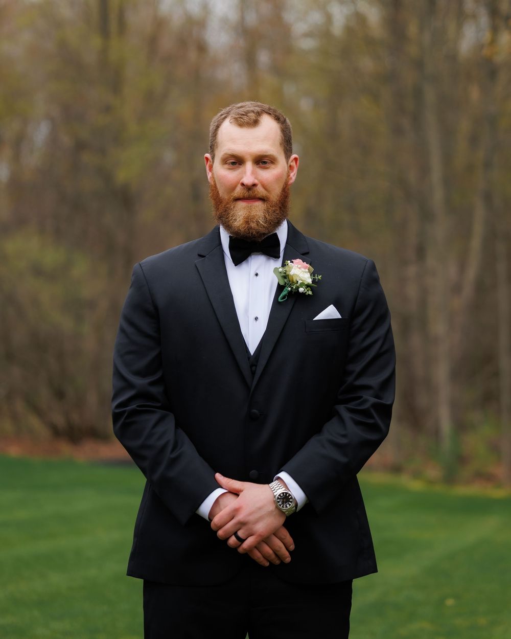 Bearded groom in black tuxedo with bow tie and boutonniere stands outdoors on green lawn with trees in background.