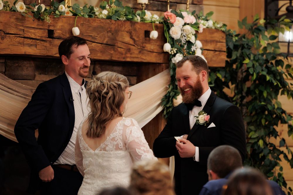 Groom smiling at bride during indoor wedding ceremony with floral arch backdrop and officiant.