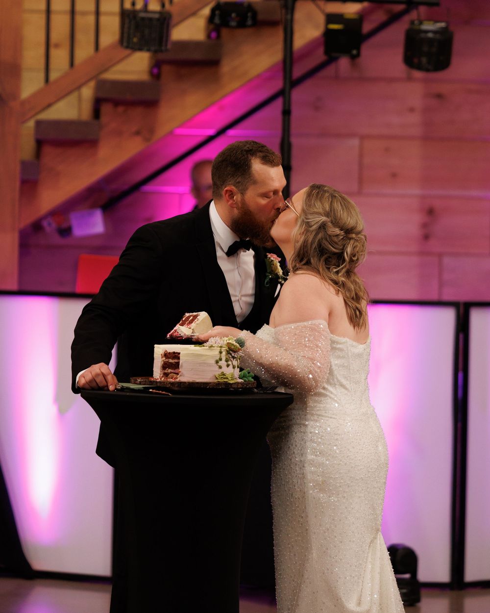 Newlyweds share a kiss during wedding cake cutting ceremony in a venue with purple uplighting and wooden staircase.