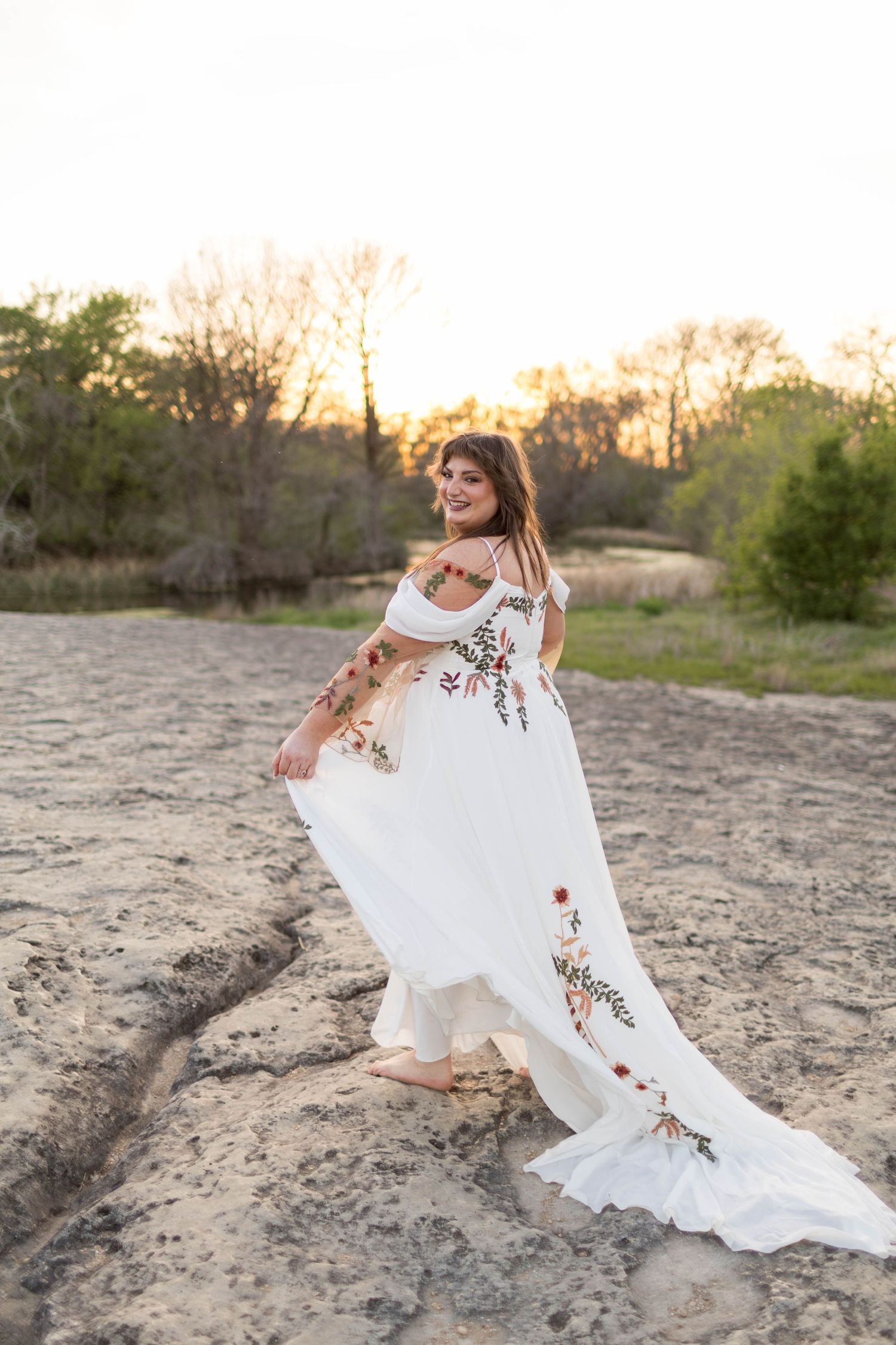 Woman in white floral embroidered boho wedding dress twirling on sandy terrain at sunset with trees in background.