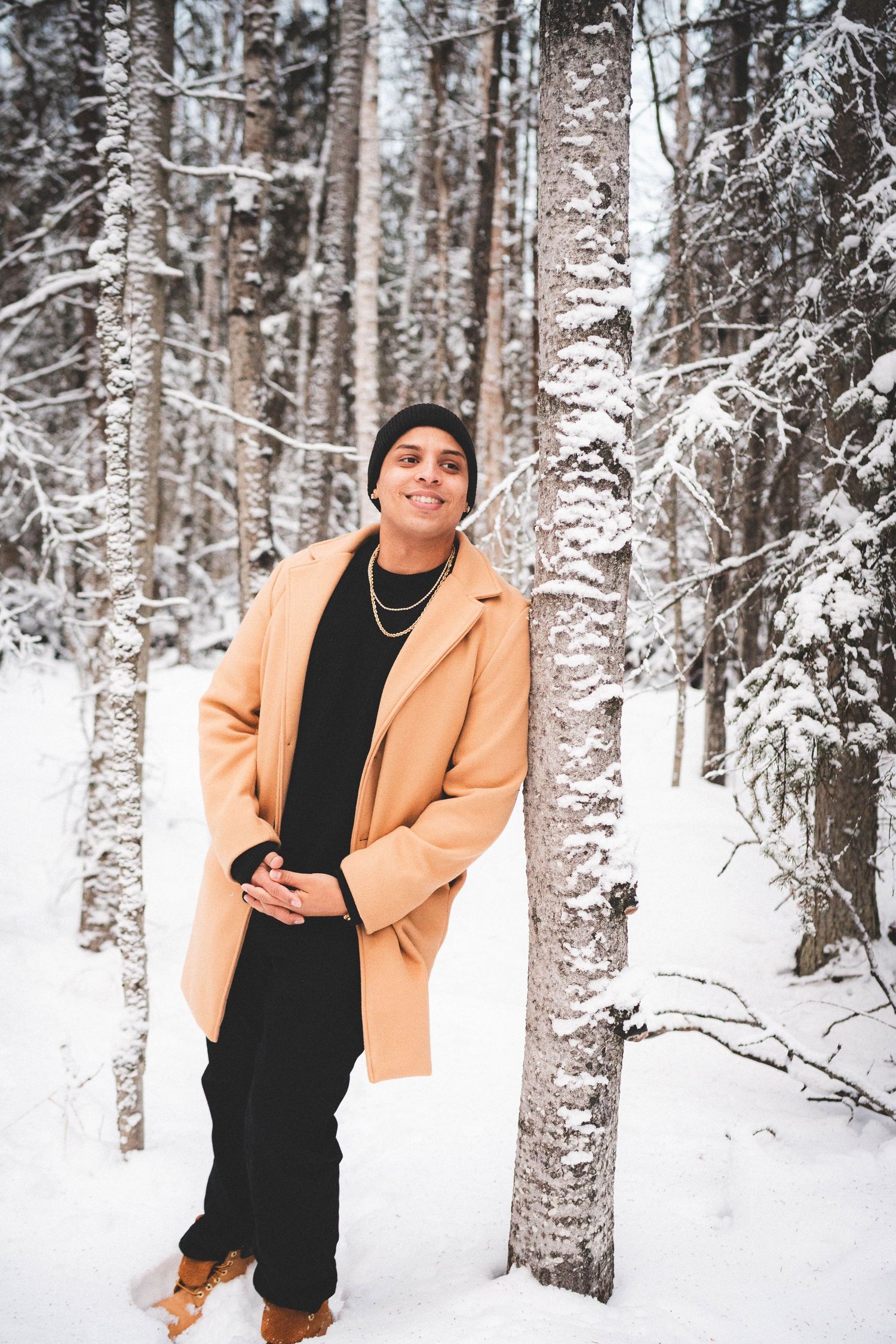 Person in beige coat and black beanie smiling in snowy winter forest, leaning against snow-covered tree.