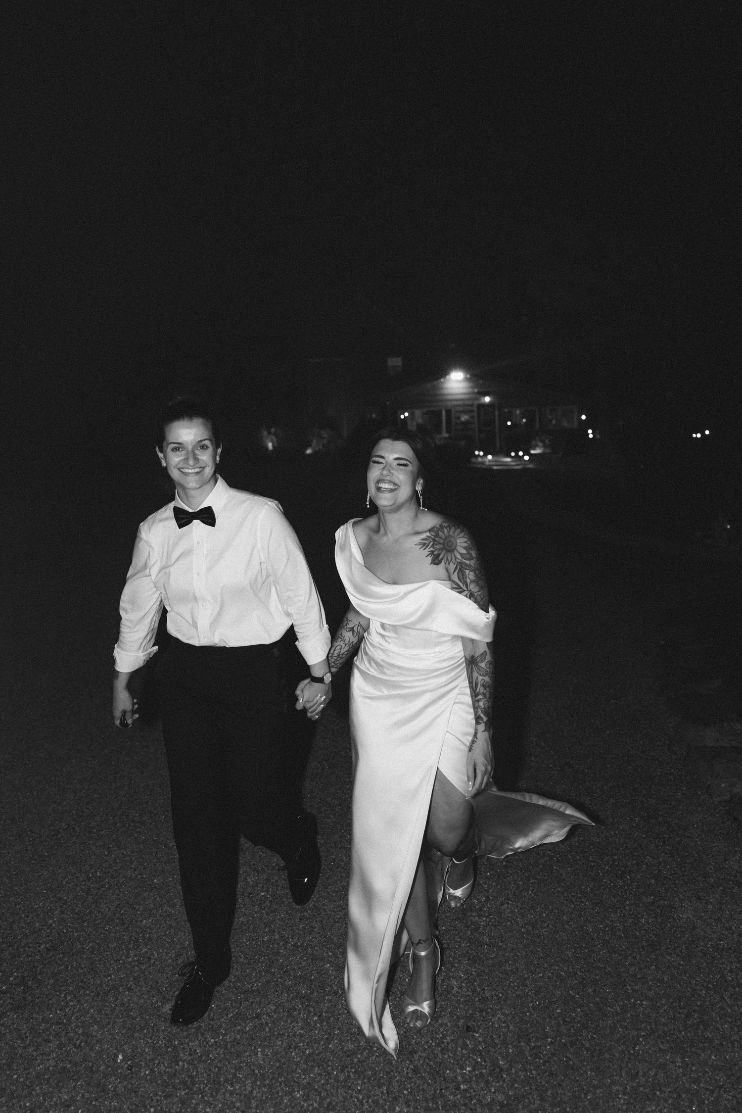 Black and white photo of a smiling couple in wedding attire holding hands and walking outdoors at night.