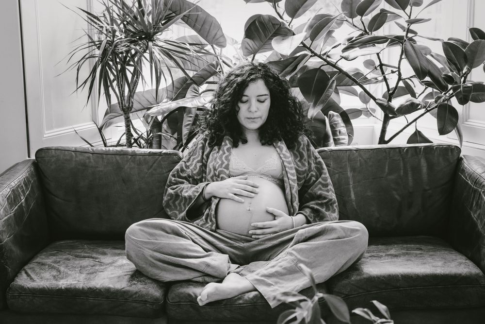 Pregnant woman sitting cross-legged on sofa cradling baby bump, surrounded by lush plants, black and white photo.