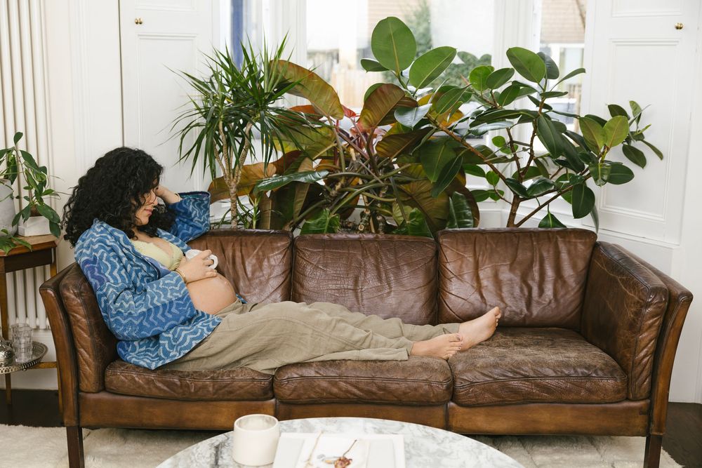 Pregnant woman relaxing on brown leather sofa surrounded by lush indoor plants in a bright living room.