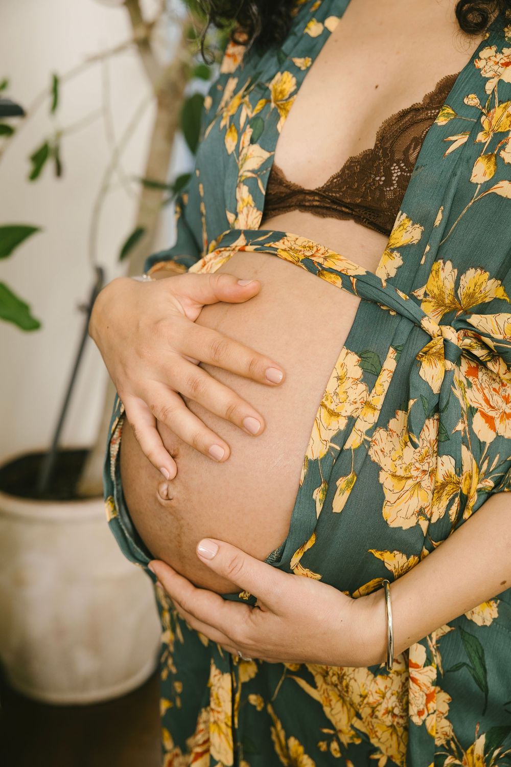 Pregnant woman in floral robe cradling bare baby bump with both hands, wearing lace bra, standing near houseplant.