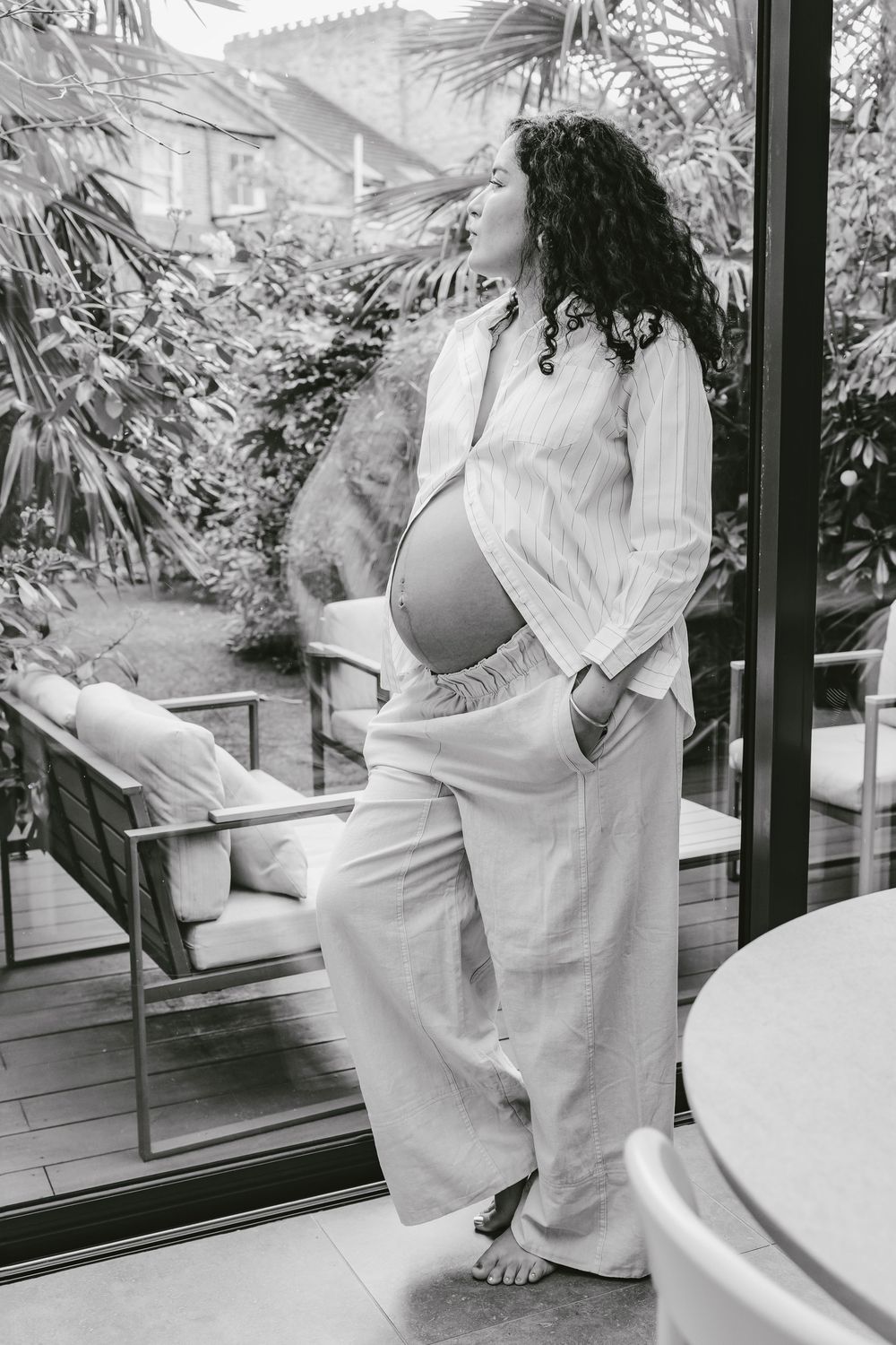 Pregnant woman in white linen outfit standing by glass doors overlooking garden.