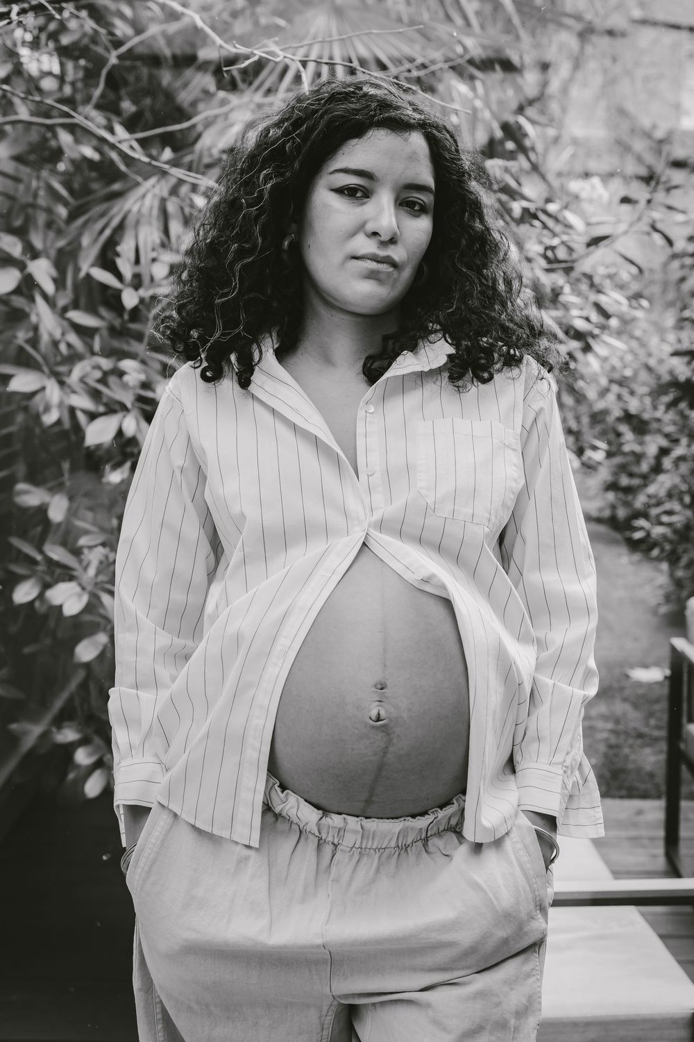 Black and white portrait of a pregnant woman with curly hair, showing her baby bump in a striped shirt outdoors.