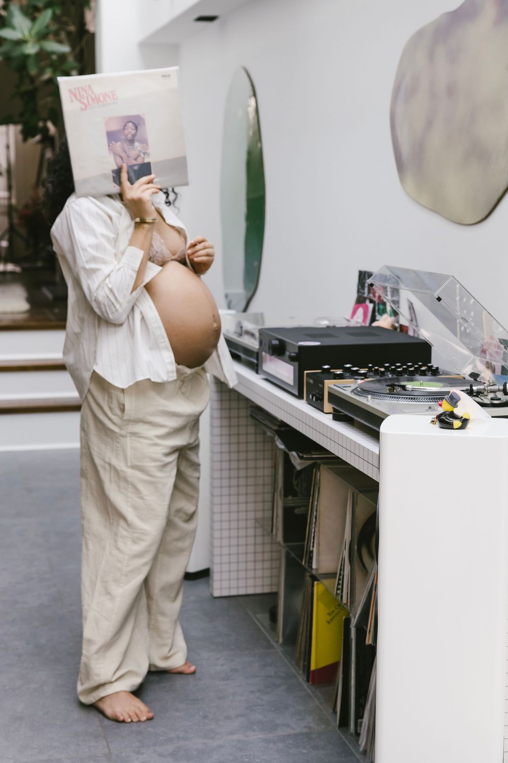 Pregnant woman holds vinyl record over face near turntable, wearing white shirt and linen pants, barefoot indoors.