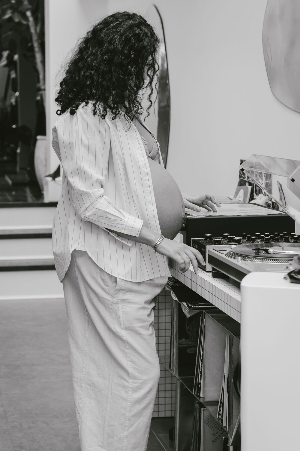 Black and white image of curly-haired pregnant woman browsing vinyl records at a turntable.