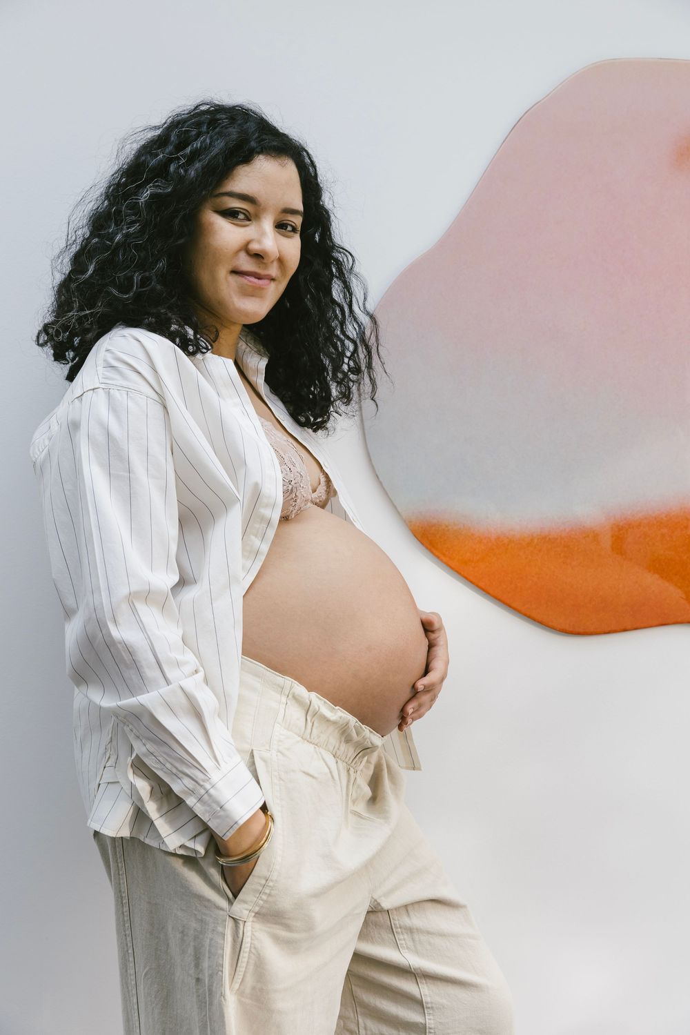 Smiling pregnant woman in white shirt holding baby bump against colorful wall art.