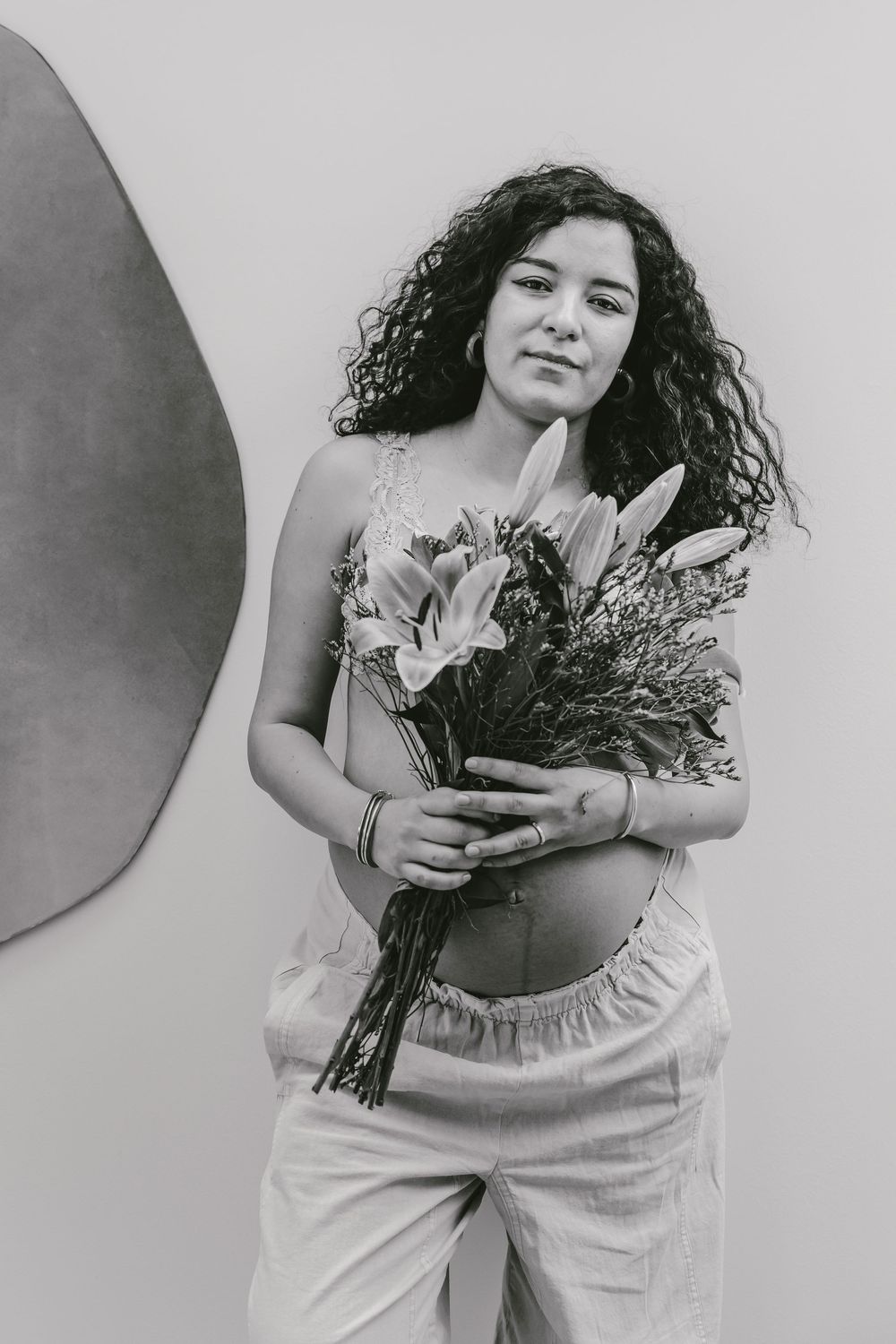 Black and white portrait of pregnant woman with curly hair holding bouquet of lilies and wildflowers indoors.