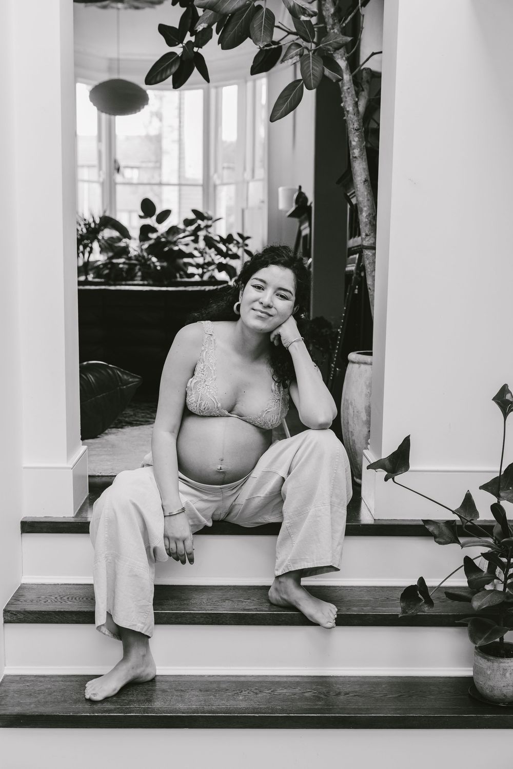Black and white photo of a smiling pregnant woman sitting on indoor steps, wearing a lace bralette and loose pants.
