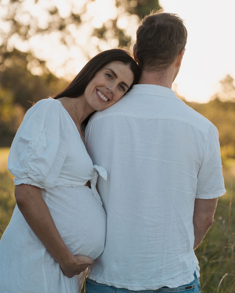 Pregnant woman in white dress smiling over her shoulder while embracing her partner in a sunlit outdoor field in rouse hill photoshoot