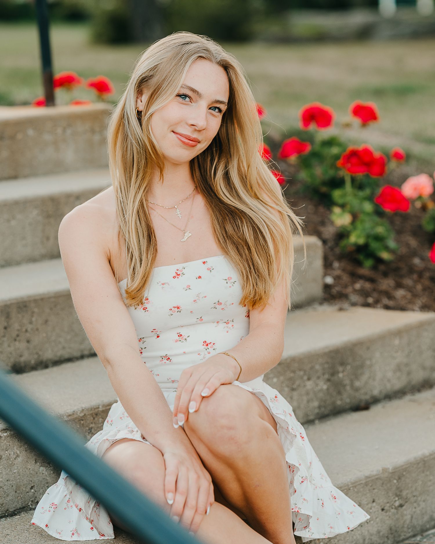 Young woman in floral sundress sitting on stone steps with red flowers in background.