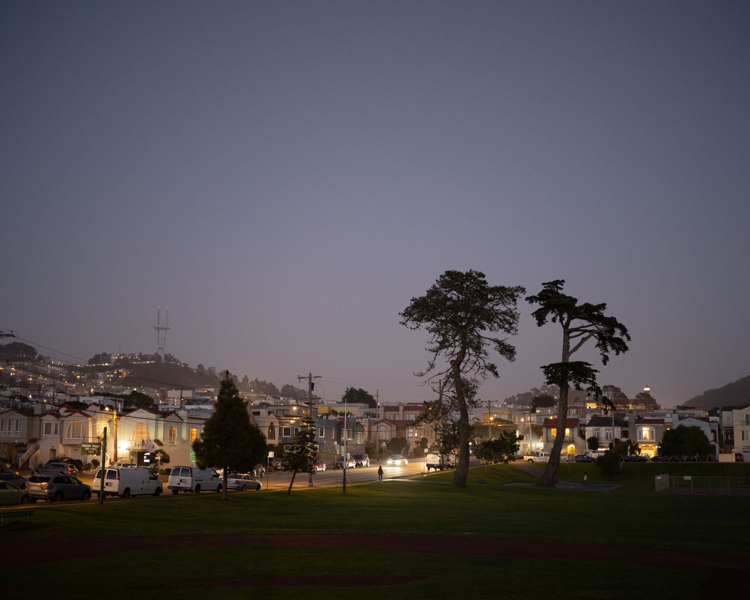 Misty San Francisco park at dusk with tall trees, green lawn, and residential buildings under overcast skies.
