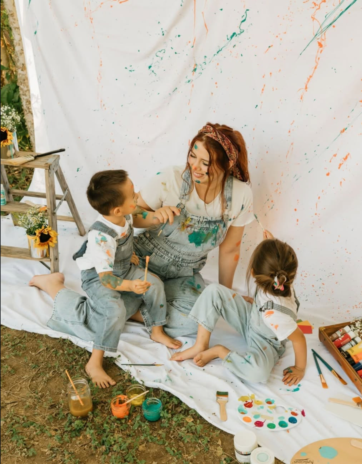 A mother and two children sit on a white sheet, joyfully painting together with colorful paints splattered on backdrop.