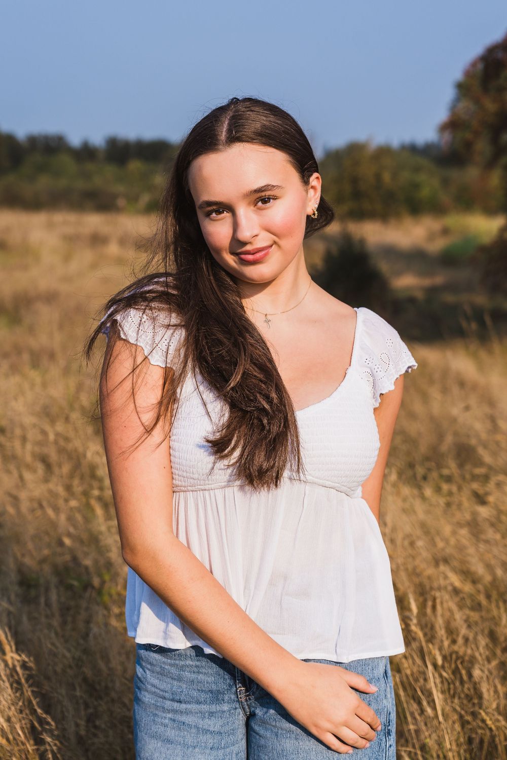 Young woman with long dark hair in white top and jeans smiling in a golden field on a sunny day.