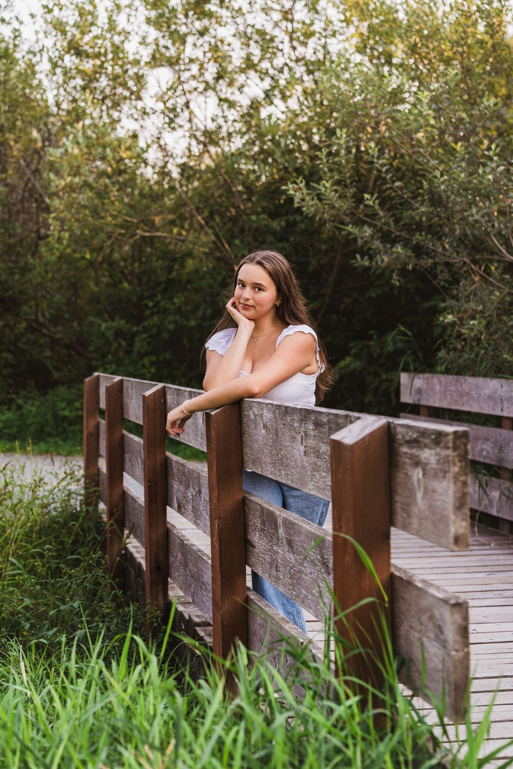 Young woman in white top leaning on wooden bridge railing surrounded by lush green trees and grass.