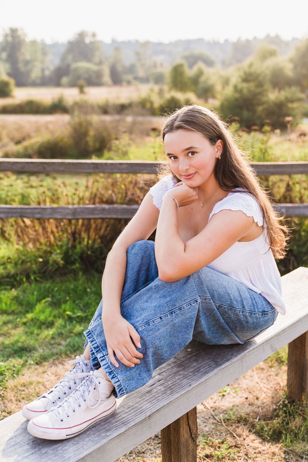 Young woman in white top and jeans sitting on wooden bench in sunny outdoor field setting.