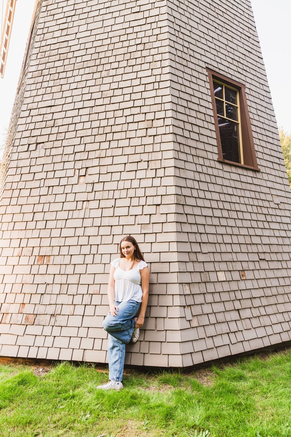 Young woman in white t-shirt and jeans leaning against a wooden shingle windmill tower on green grass.