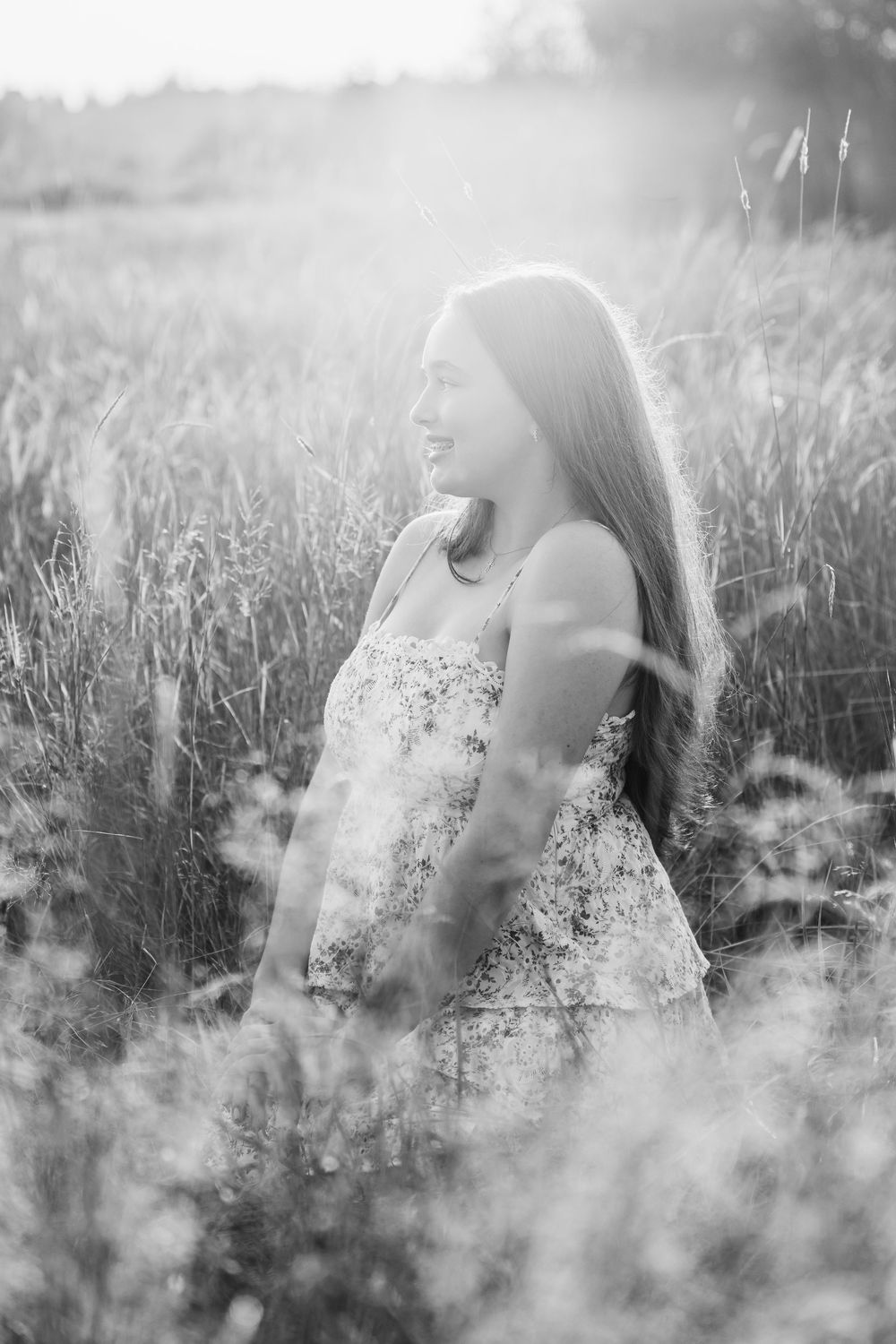 Black and white portrait of a young woman smiling in a sunlit field of tall grass wearing a floral dress.