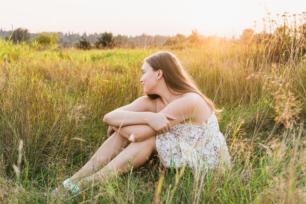 Young woman in floral dress sitting in golden meadow at sunset, hugging knees while gazing peacefully into distance.