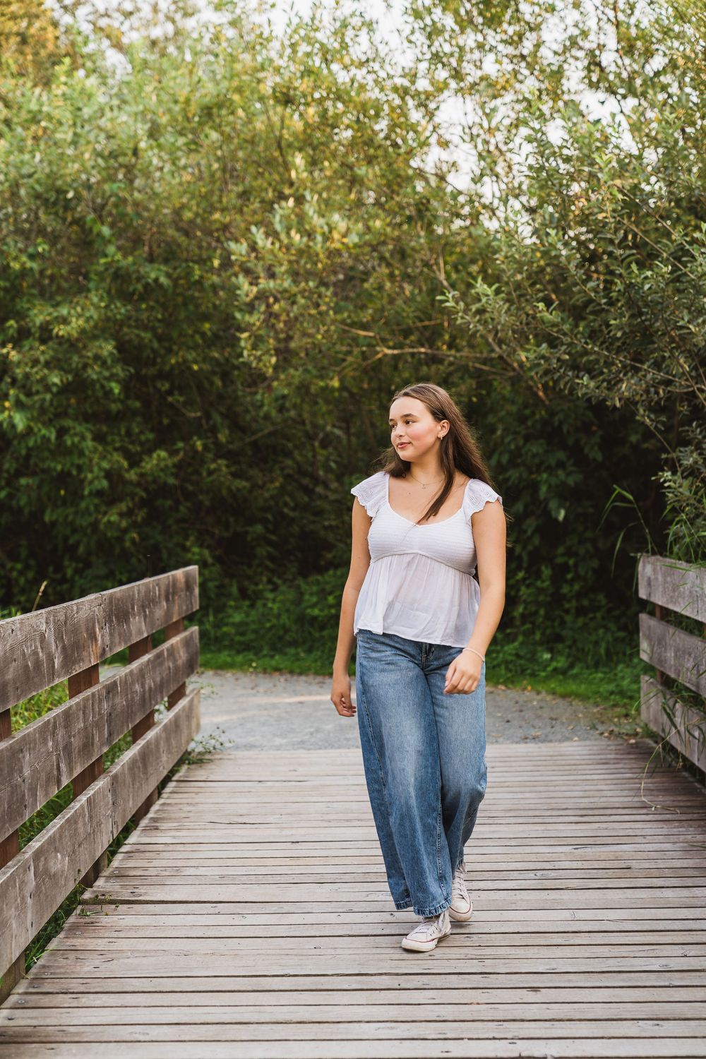 Young woman in white top and wide-leg jeans walking on wooden boardwalk path surrounded by lush green trees.