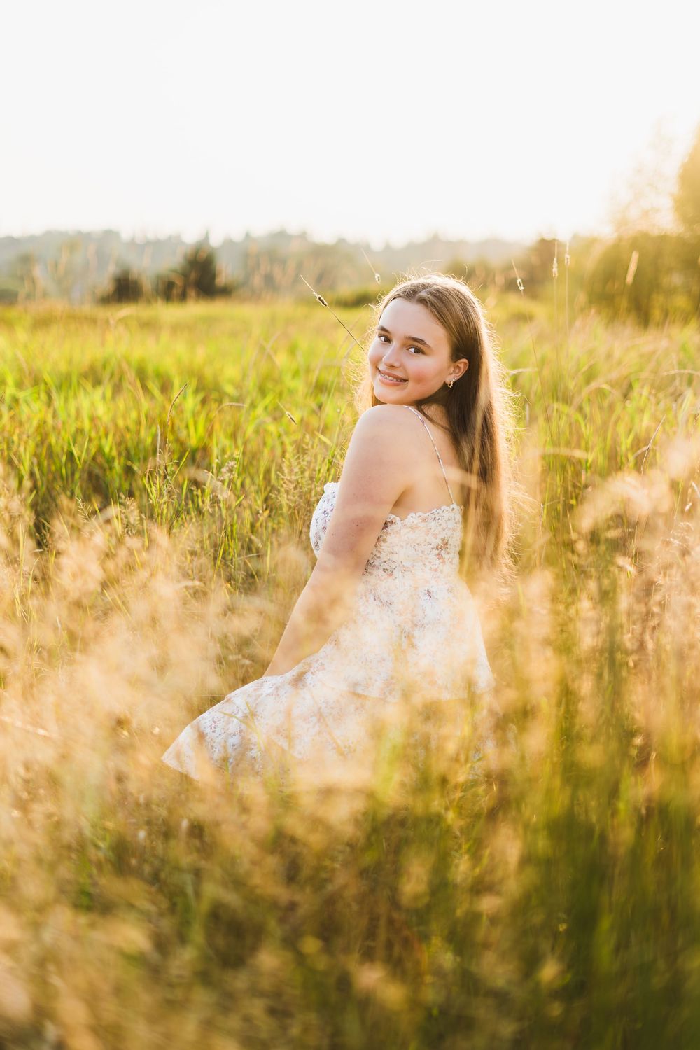 Young woman in floral dress smiling over shoulder in golden sunlit meadow with tall grass during sunset.