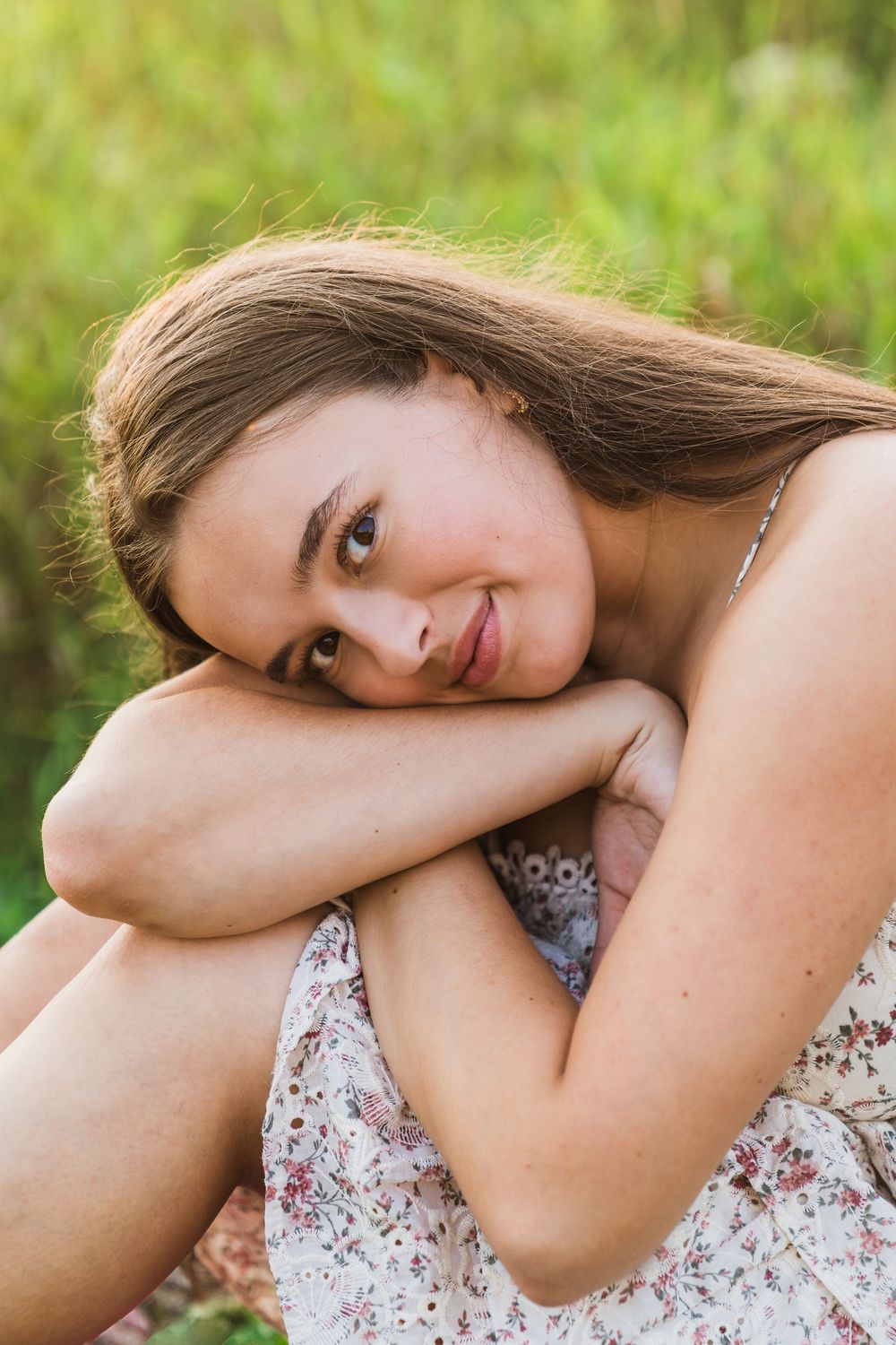 Young woman in floral dress sitting in green field, resting head on knees, looking at camera.