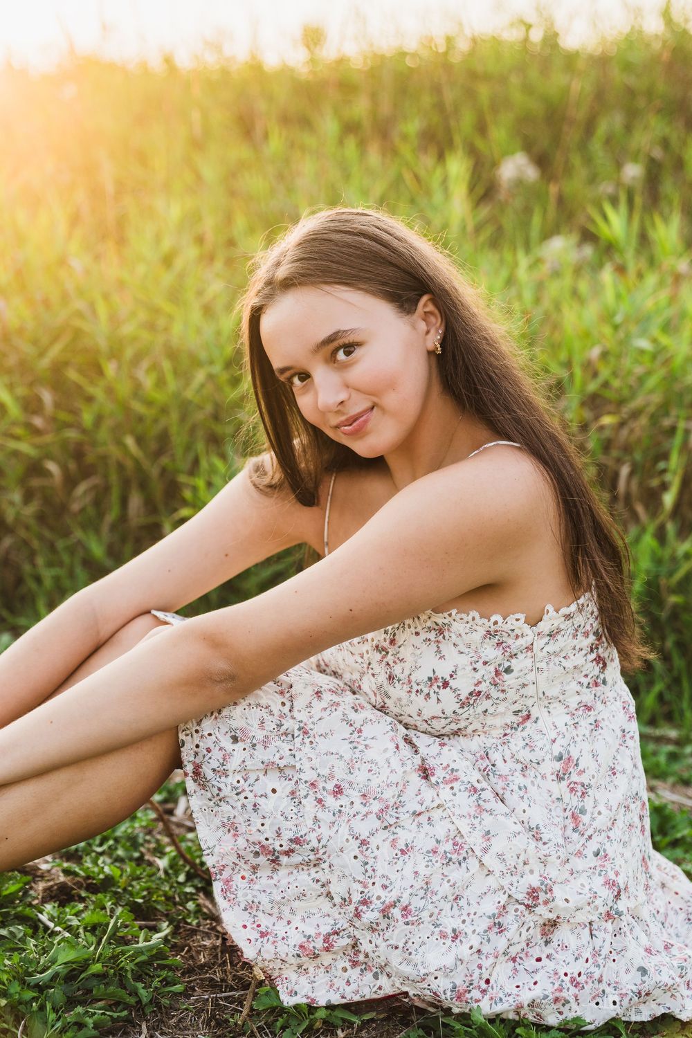 Young woman in floral sundress sitting in green meadow grass smiling in warm golden sunset light.