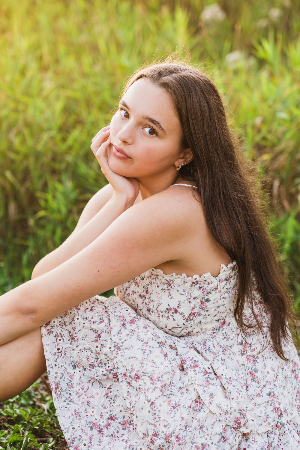 Young woman in floral sundress sitting in green grass field, posing thoughtfully with hand on chin in golden sunlight.