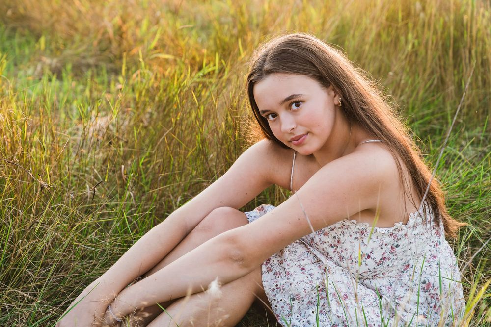 Young woman in floral dress sitting in golden grass field during sunset, looking over shoulder.
