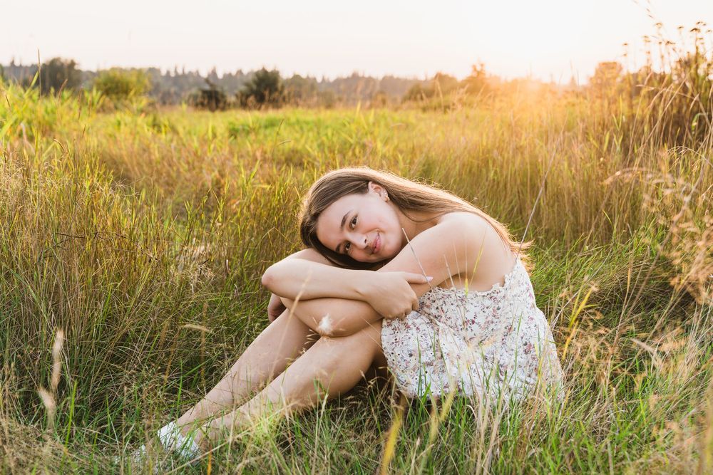 Young woman in floral dress sitting peacefully in a sunlit meadow, hugging her knees among tall green grasses.