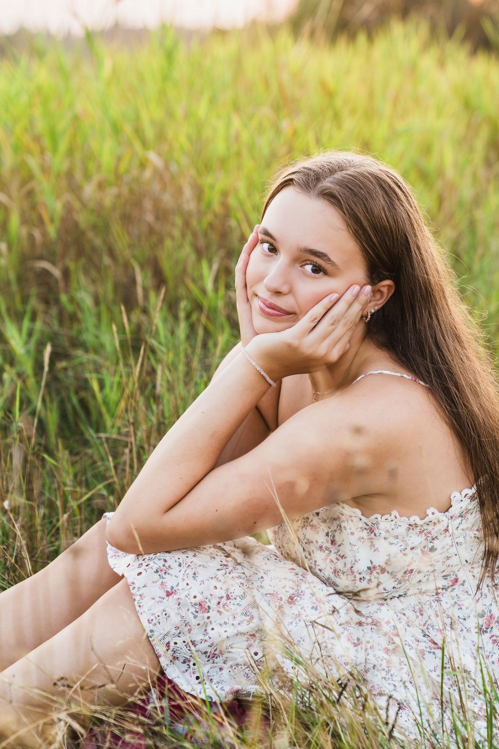 Young woman in floral sundress sitting in tall grass field, resting cheek on hand, smiling softly.