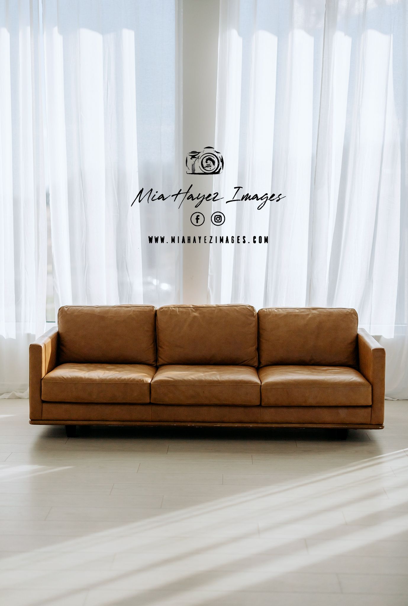 Modern tan leather three-seat sofa in a bright minimalist room with sheer white curtains and light hardwood floors.