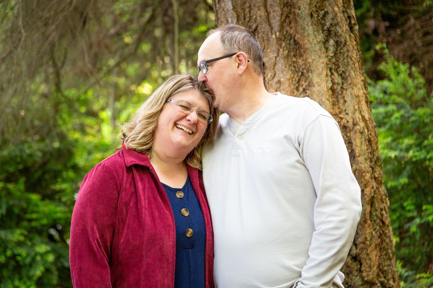 A couple shares a tender moment outdoors, man kissing woman's head near a tree, both smiling in a lush green forest.