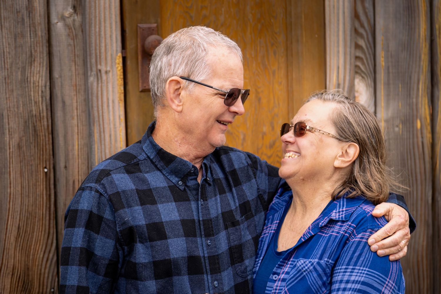 Smiling senior couple in sunglasses sharing a warm embrace against a rustic wooden backdrop.