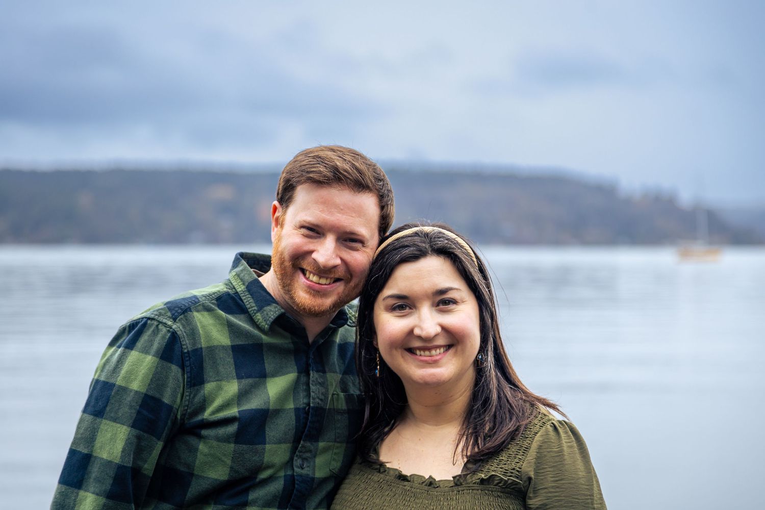 Smiling couple posing outdoors near a lake with scenic hills in the background on a cloudy day.