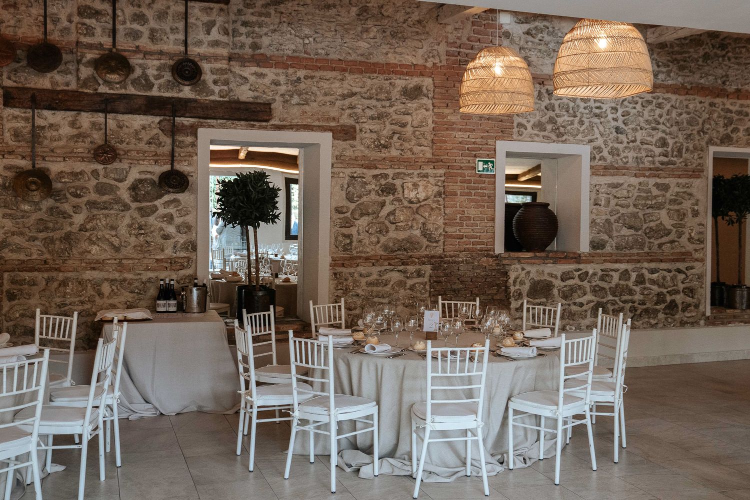 Elegant rustic wedding venue with white Chiavari chairs, round tables, stone walls, and woven pendant lights.