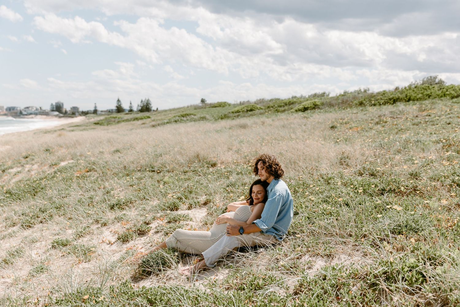 A person sits on a grassy hillside overlooking a scenic landscape during a cloudy day.