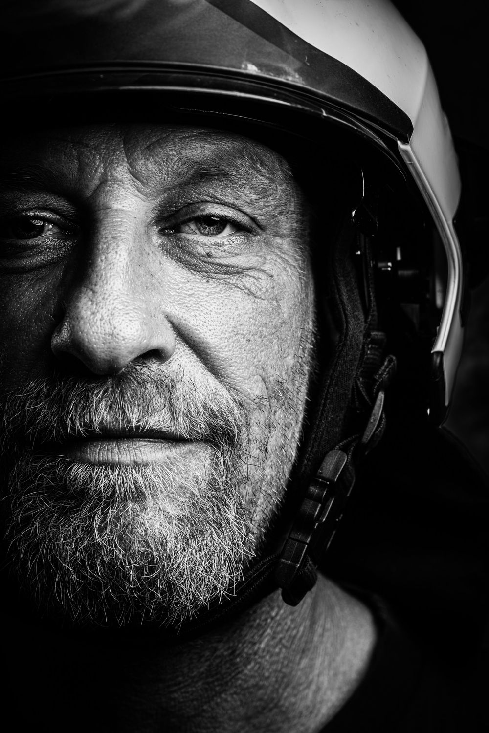 Striking black and white close-up portrait of weathered bearded firefighter wearing white helmet.