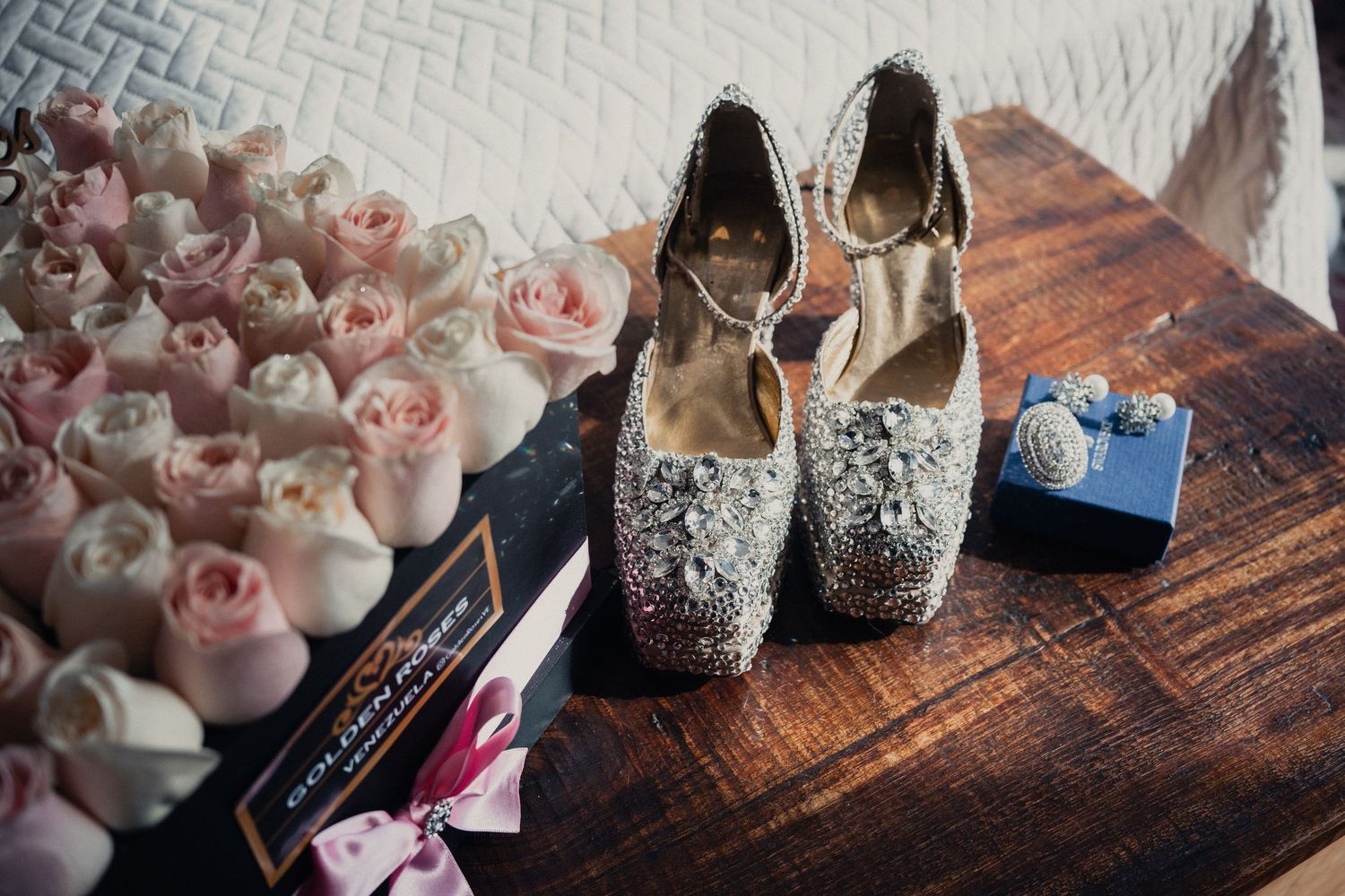 Wedding details including sparkly high heels, delicate pink roses, and makeup accessories arranged on a rustic wooden surface.
