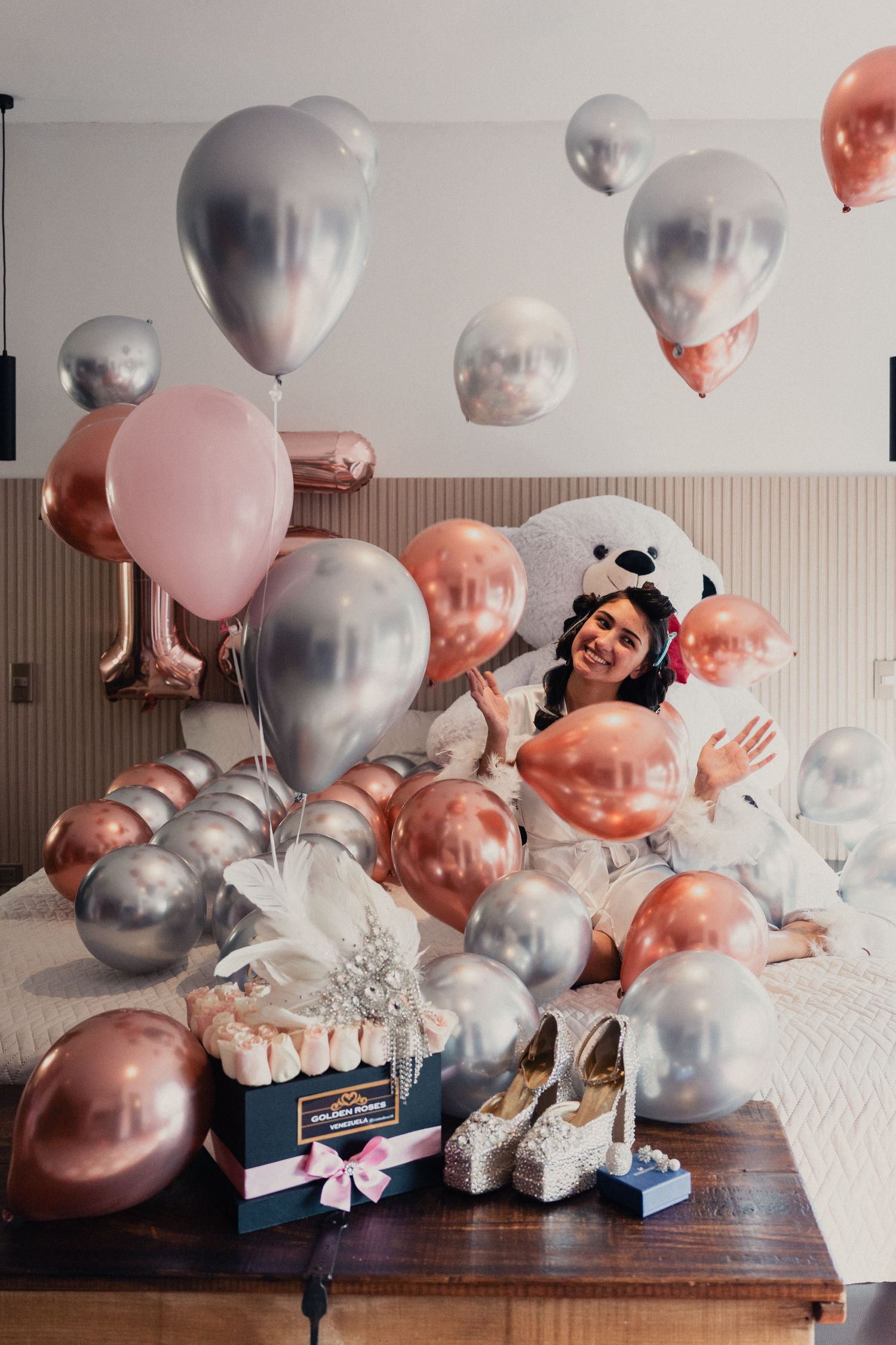 Festive celebration scene with silver and rose gold balloons surrounding a gift box and elegant shoes on wooden table.