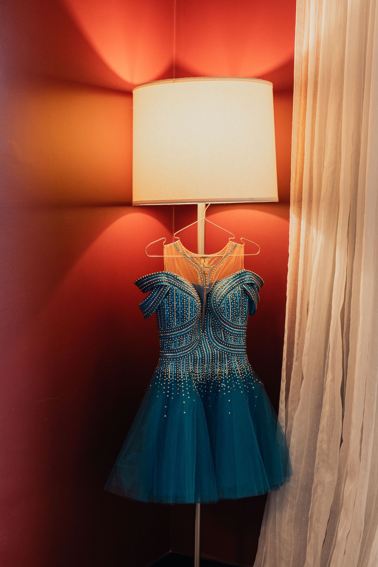 A sparkly blue formal dress with beaded bodice and tulle skirt hangs illuminated by a white lamp shade in a red corner.