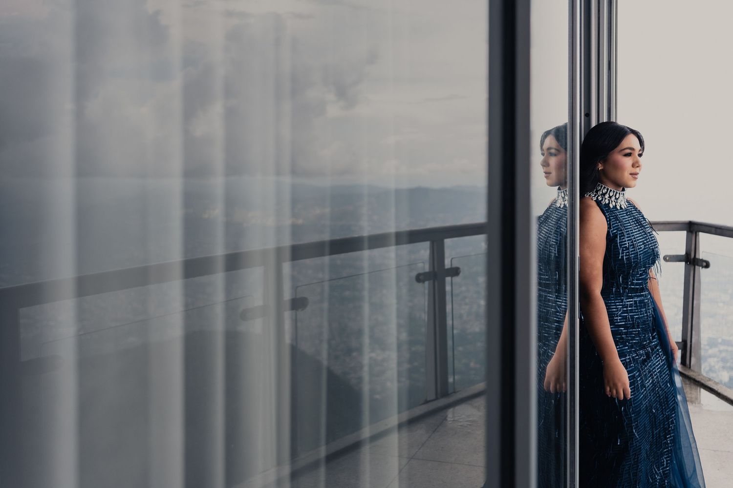 Woman in navy blue dress stands thoughtfully by modern glass windows in an elegant interior space.