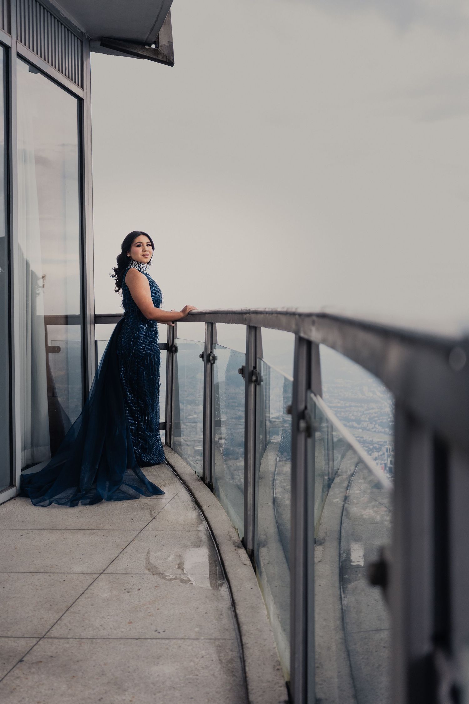 A person in a dark dress sits contemplatively on a high-rise balcony overlooking a cityscape at dusk.