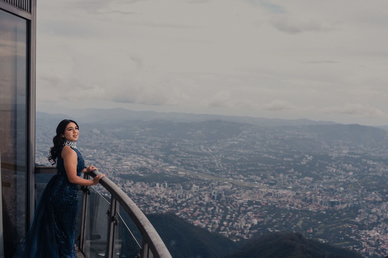 A person in a blue dress admires the sweeping cityscape view from a high observation deck on a cloudy day.