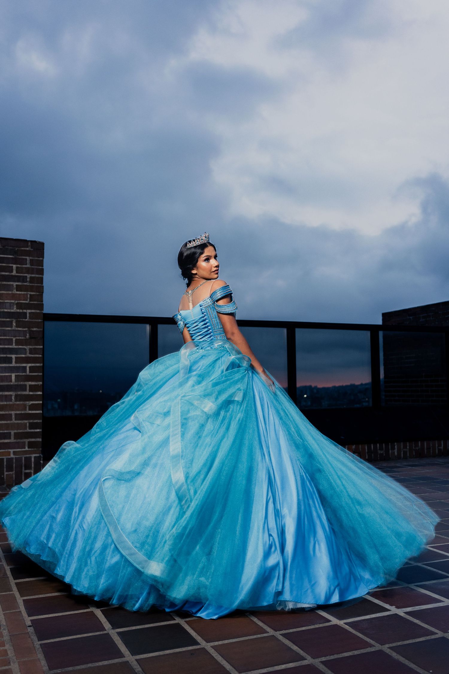 A stunning turquoise quinceañera ball gown with layers of tulle fabric photographed against a cloudy sky on a rooftop.