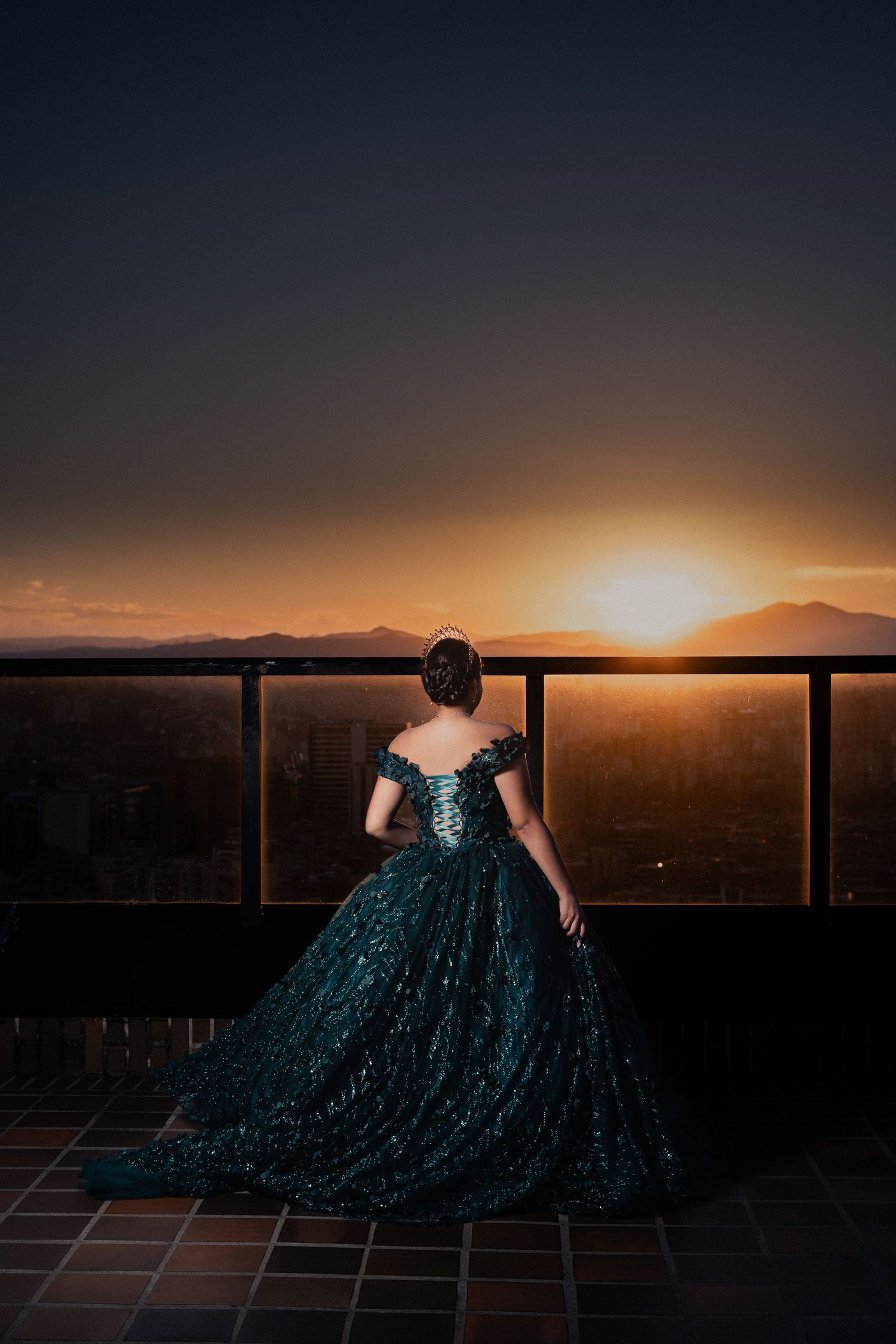 A figure in a sparkling teal evening gown poses against a dramatic sunset backdrop over mountains and desert landscape.