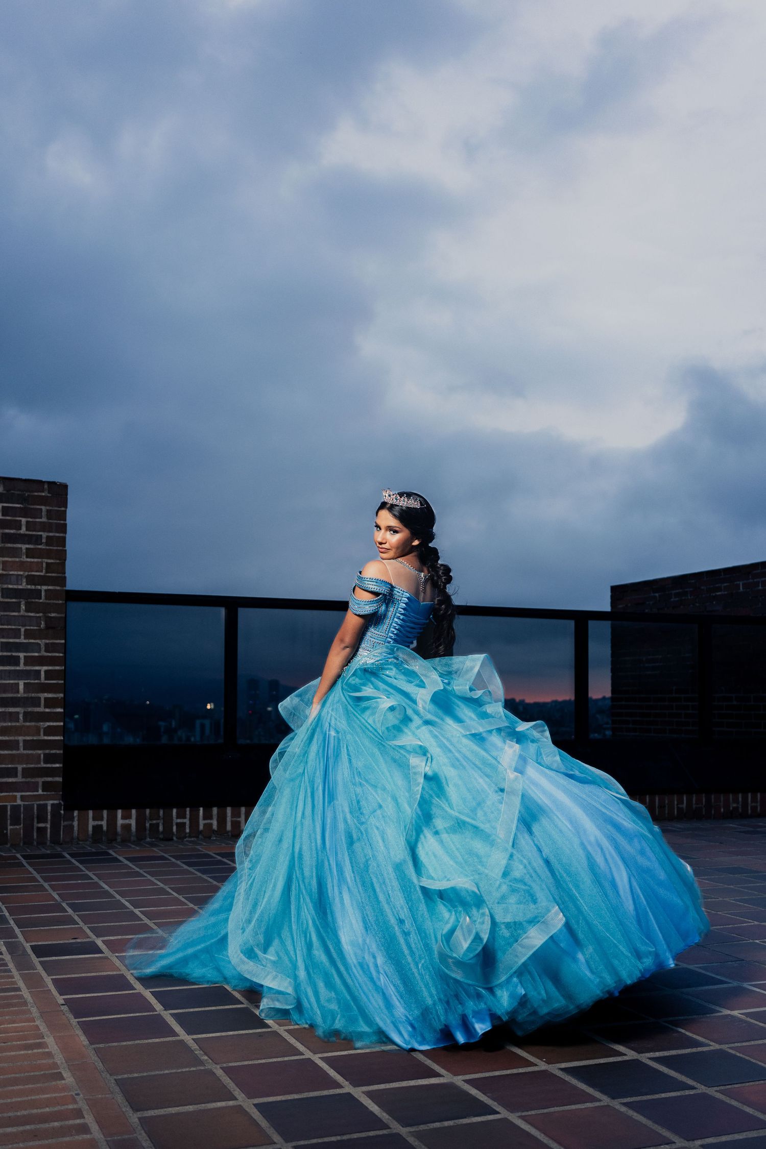A person in a flowing turquoise quinceañera gown poses dramatically on a rooftop against a cloudy sky.